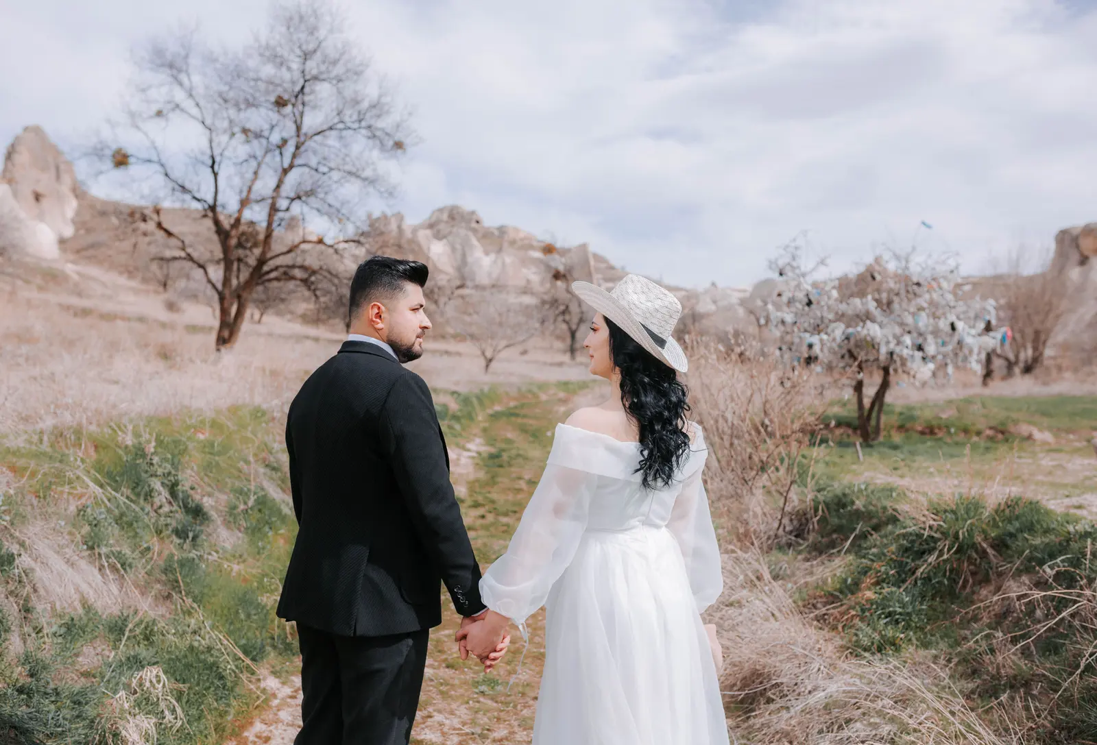 A bride in a white hat and her groom walking hand in hand along a trail between rock formations