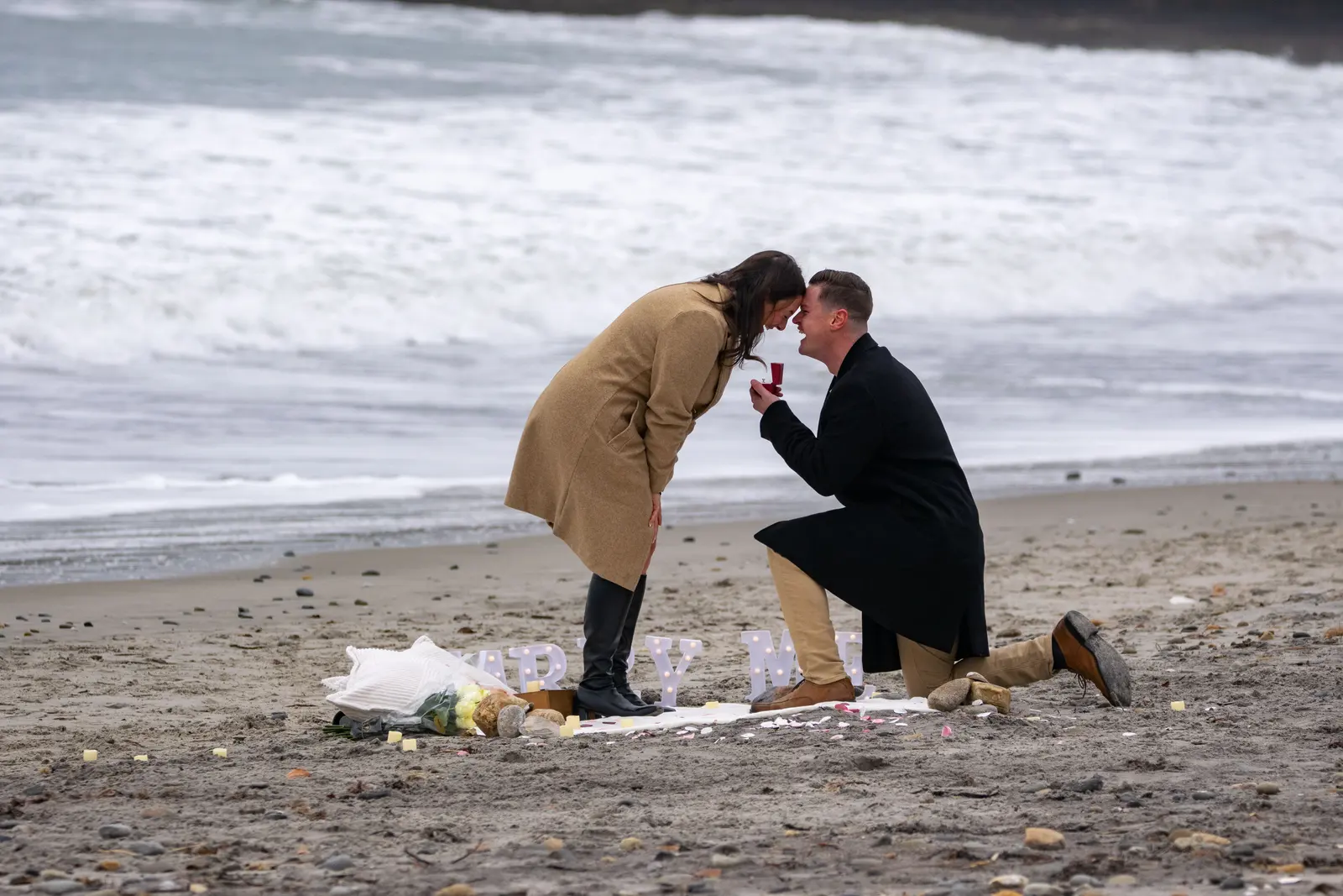 A man on one knee holding out a ring to his partner on a beach