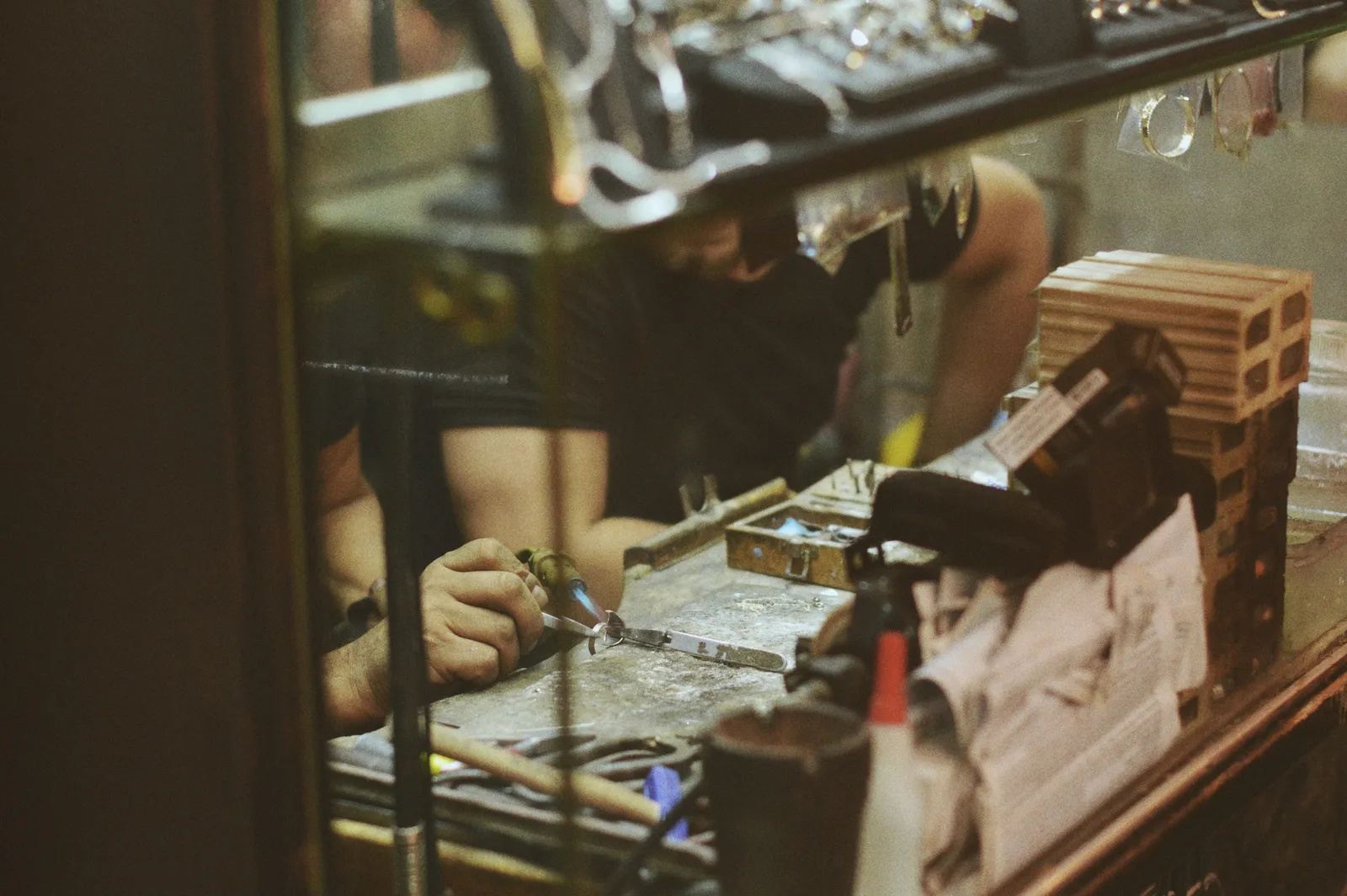 A jeweller working at their bench with a torch, seen through the shop window