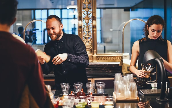 A bartender laughing with a guest while a colleague pours drinks behind a busy bar