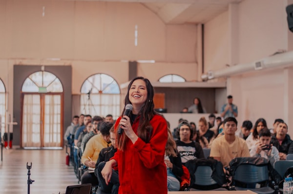 A smiling woman in a red jacket takes the microphone in front of a full hall