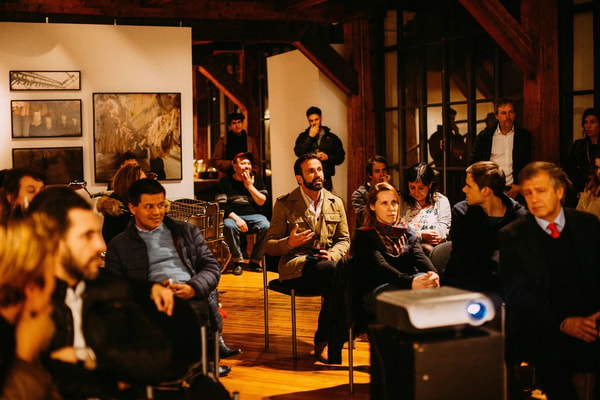A small audience in a timber-beamed gallery, one guest asking a question beside the projector