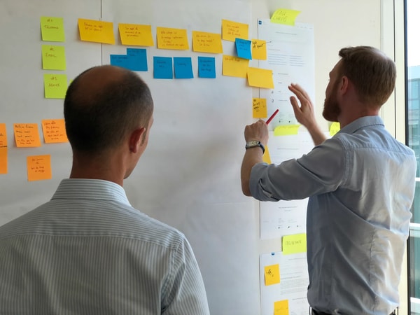 Two men at a whiteboard wall, one adding a sticky note to a row of yellow and blue ones