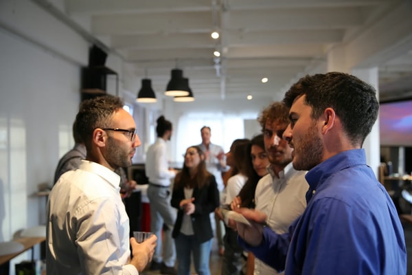 Two men talking over drinks at a daytime reception, the room busy behind them