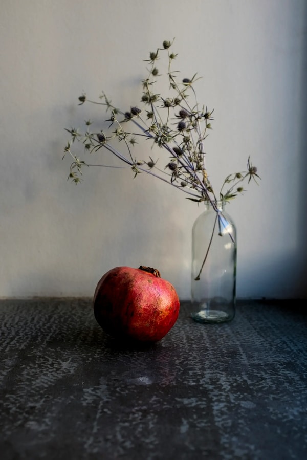 Still life of a pomegranate beside dried thistle stems in a glass bottle