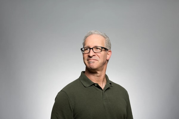 Grey-haired man in glasses and an olive polo shirt against a plain grey studio backdrop