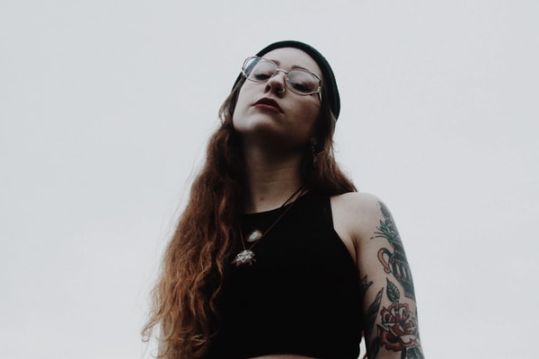 Woman in glasses and a black beanie, tattooed arm, shot from below against a pale sky