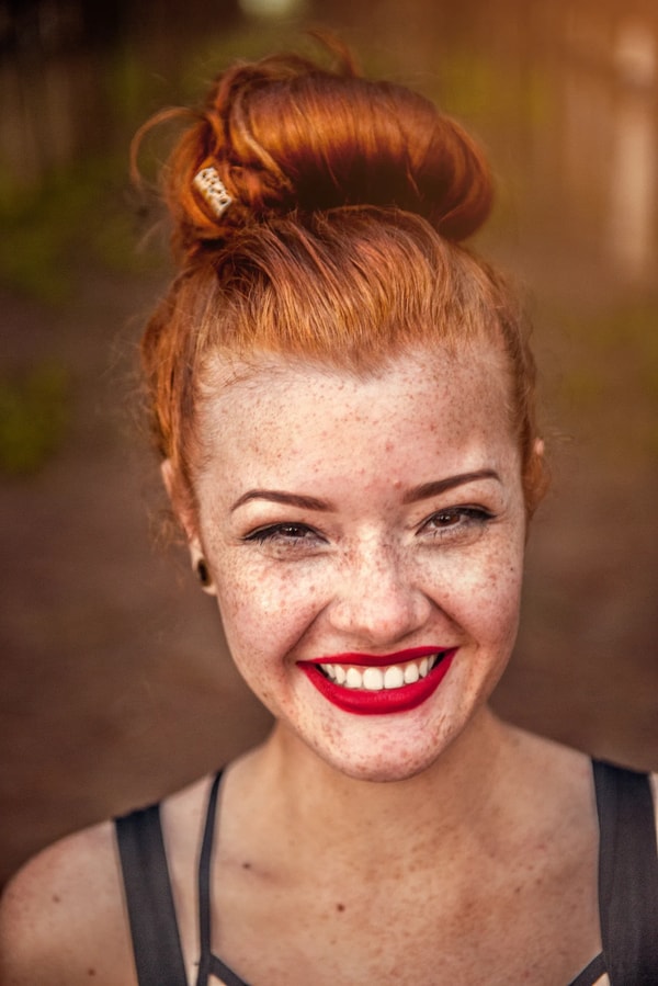 Red-haired woman with freckles and red lipstick smiling broadly, hair in a top knot