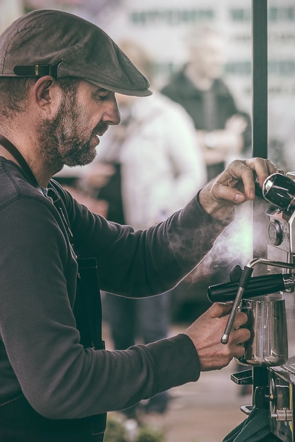 Barista in a flat cap steaming milk at an espresso machine