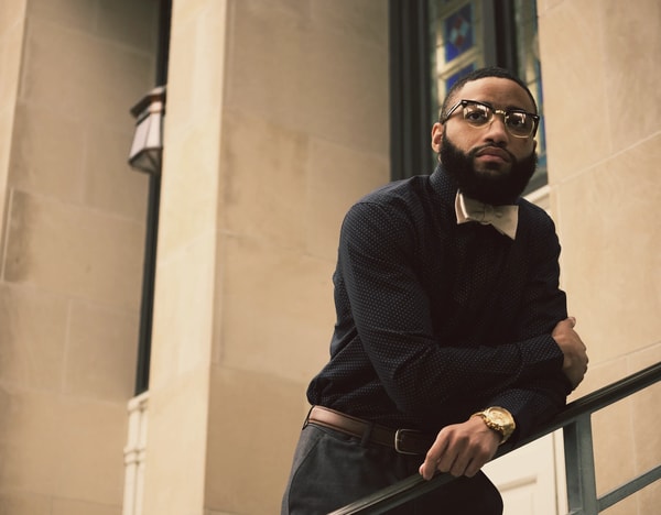 Man with a beard and glasses leaning on a stair rail outside a stone building