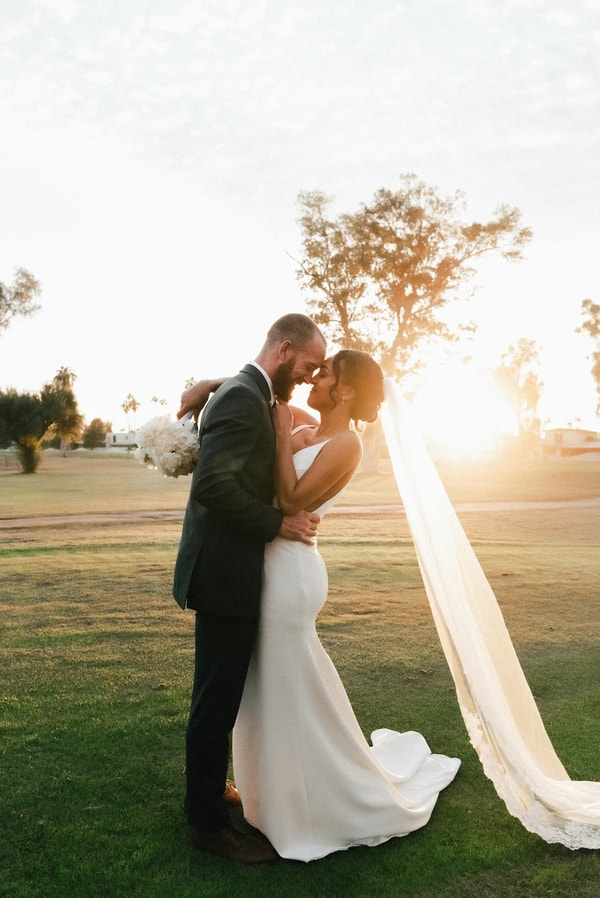 Couple embracing on a lawn at sunset, the bride’s train sweeping over the grass