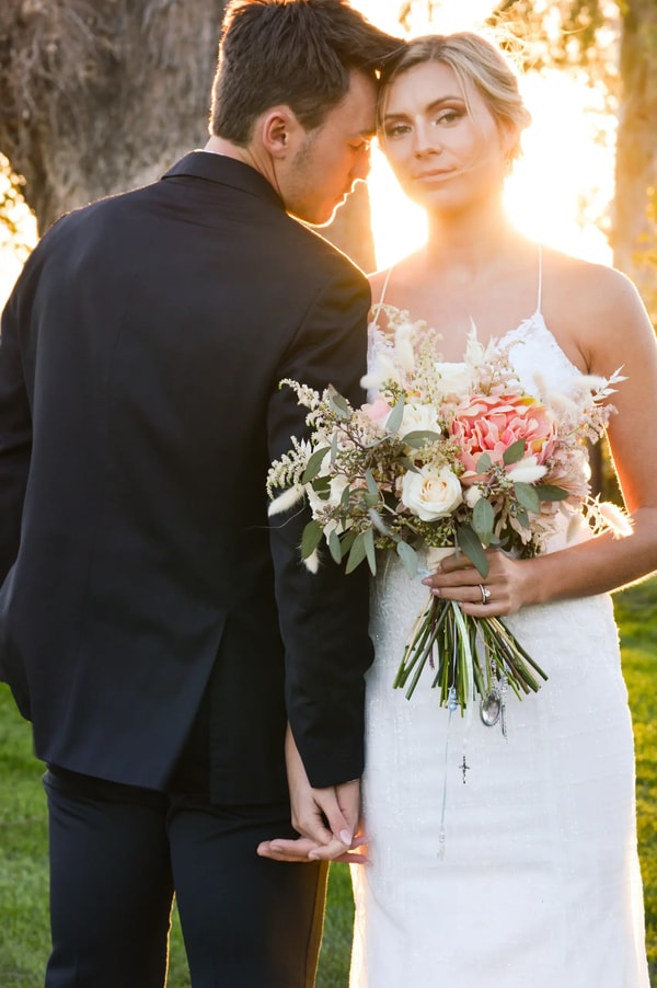 Bride holding a peach and cream bouquet, leaning into the groom at sunset