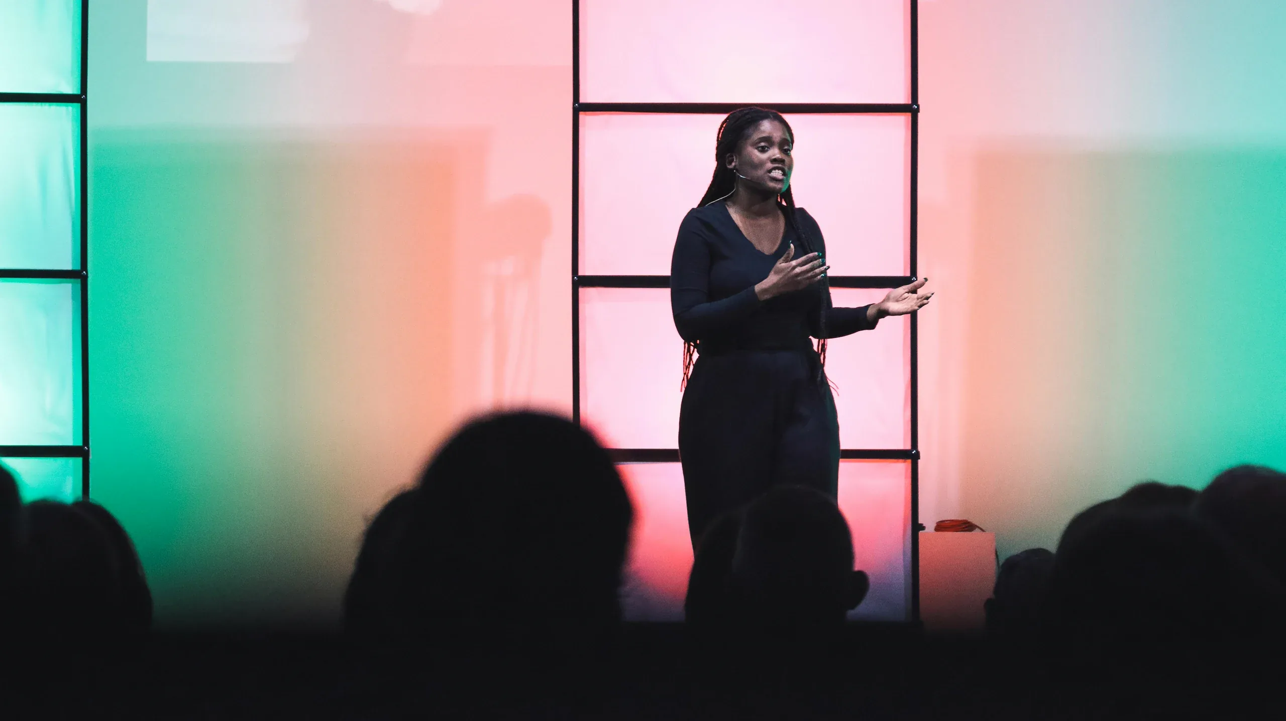 A speaker mid-sentence on a stage lit pink and green, the audience in silhouette below