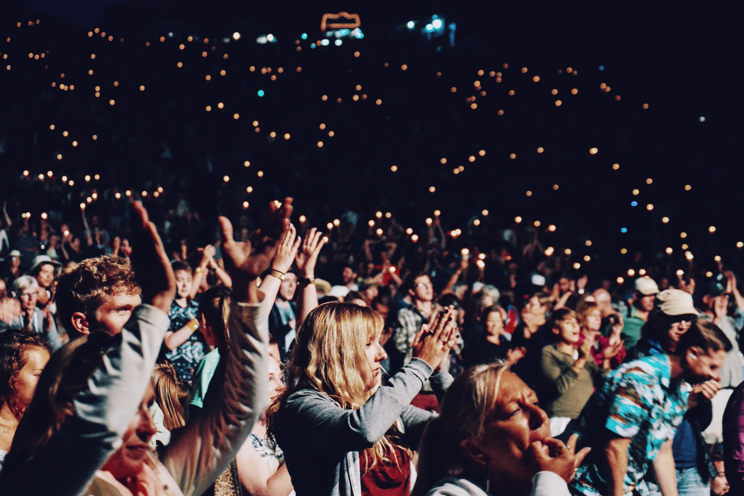 A crowd cheering and singing along with arms raised, hundreds of small lights behind them