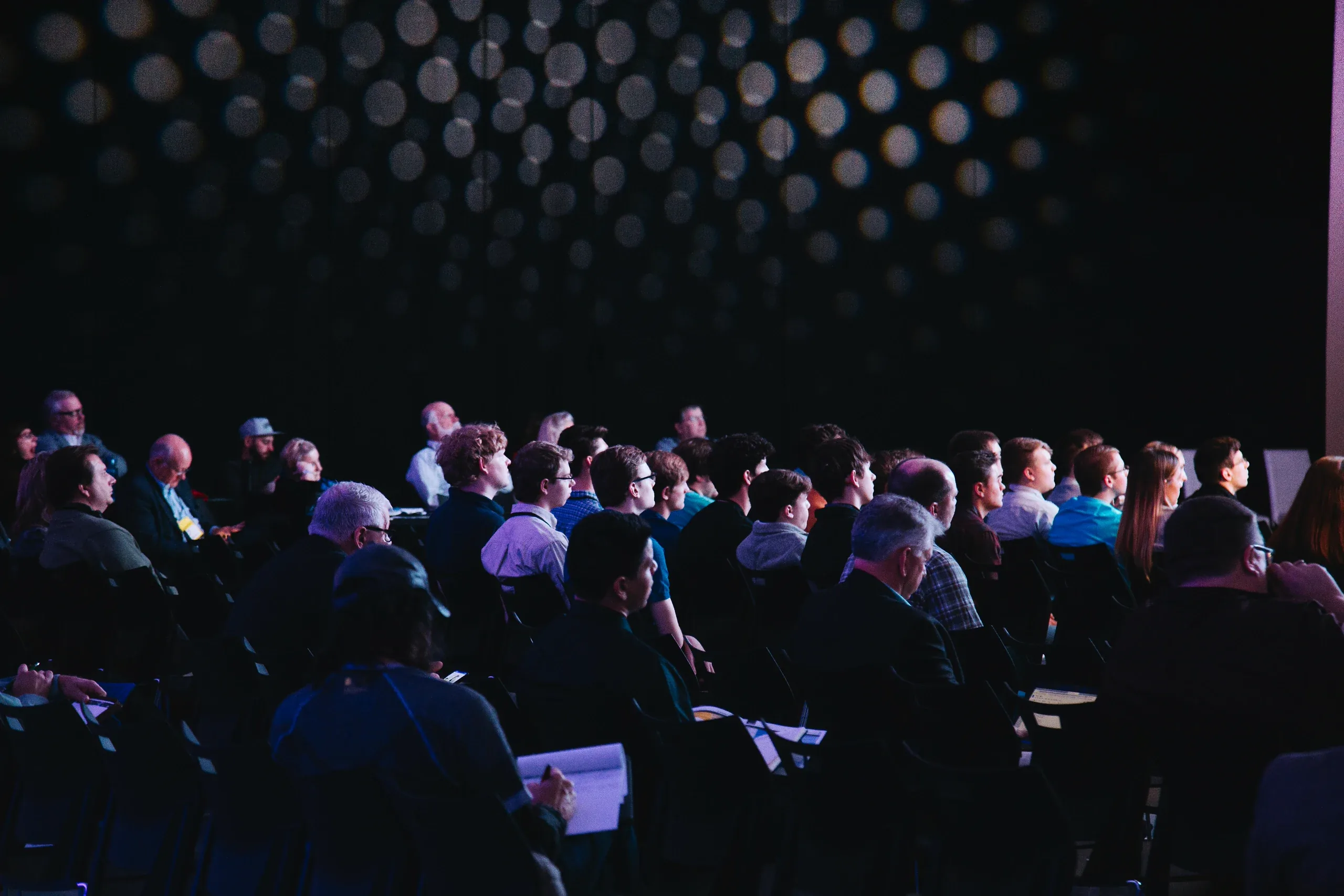 A seated conference audience in a dark hall, turned towards the stage, notebooks on their knees