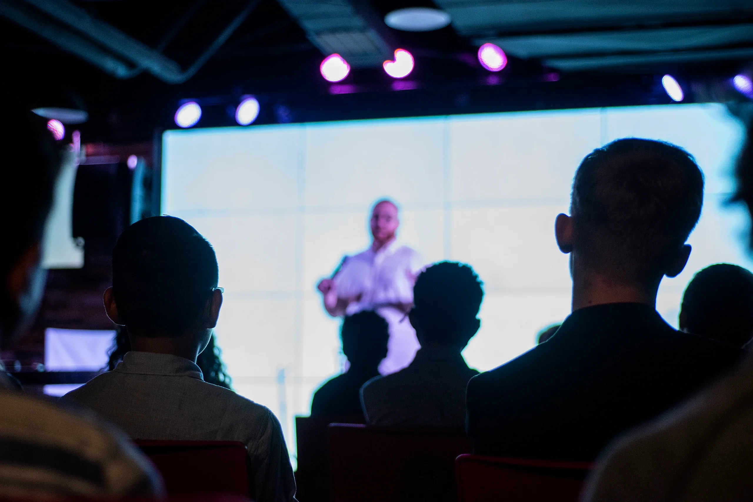 A speaker in front of a bright screen, seen past the backlit heads of the audience