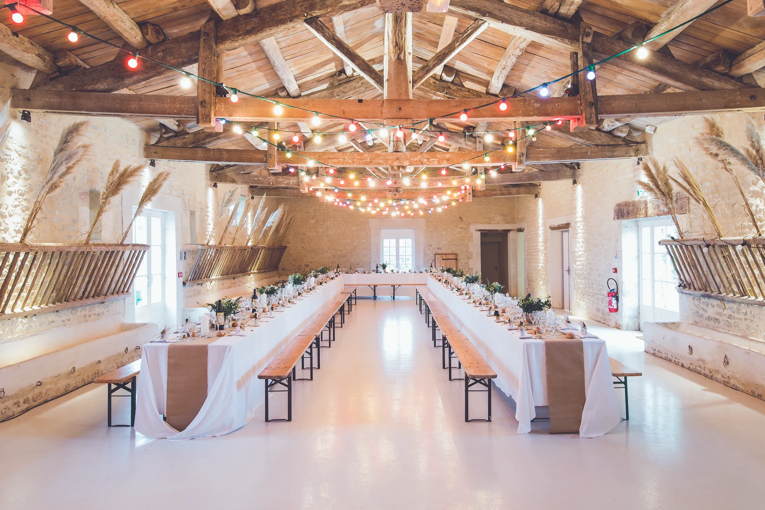 Long dinner tables set under festoon lights in a whitewashed hall with timber beams