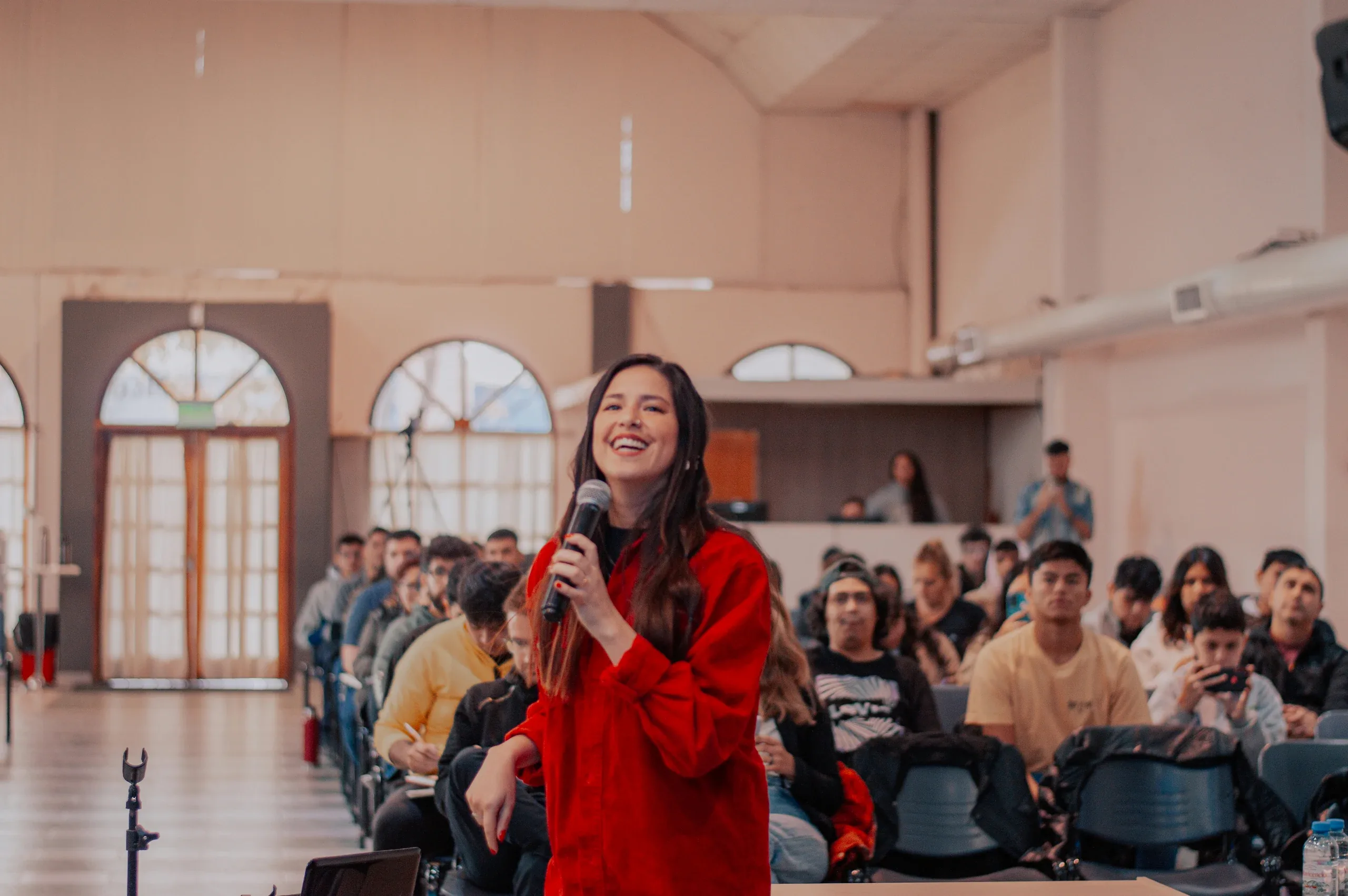A smiling woman in a red jacket takes the microphone in front of a full hall