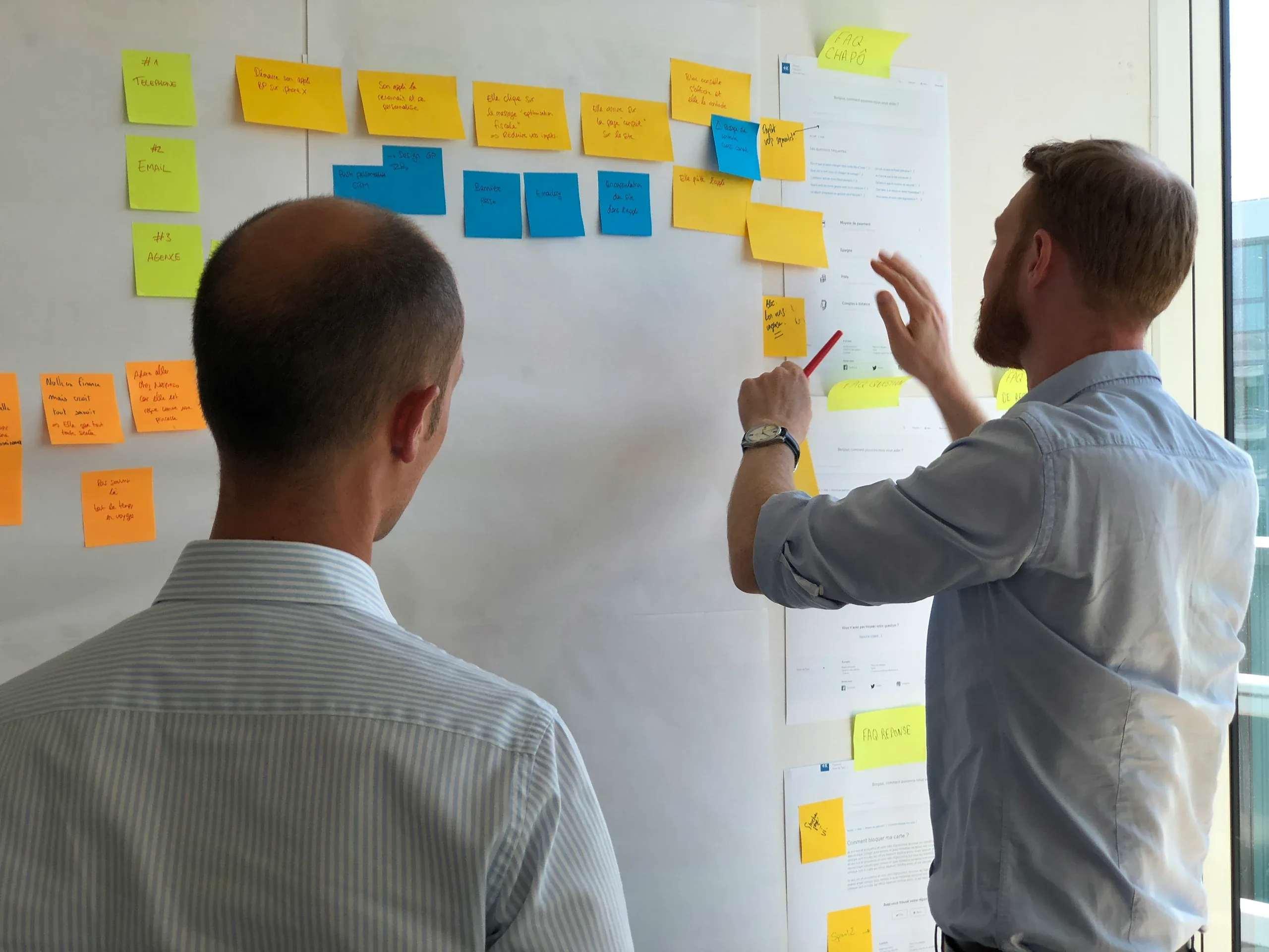 Two men at a whiteboard wall, one adding a sticky note to a row of yellow and blue ones