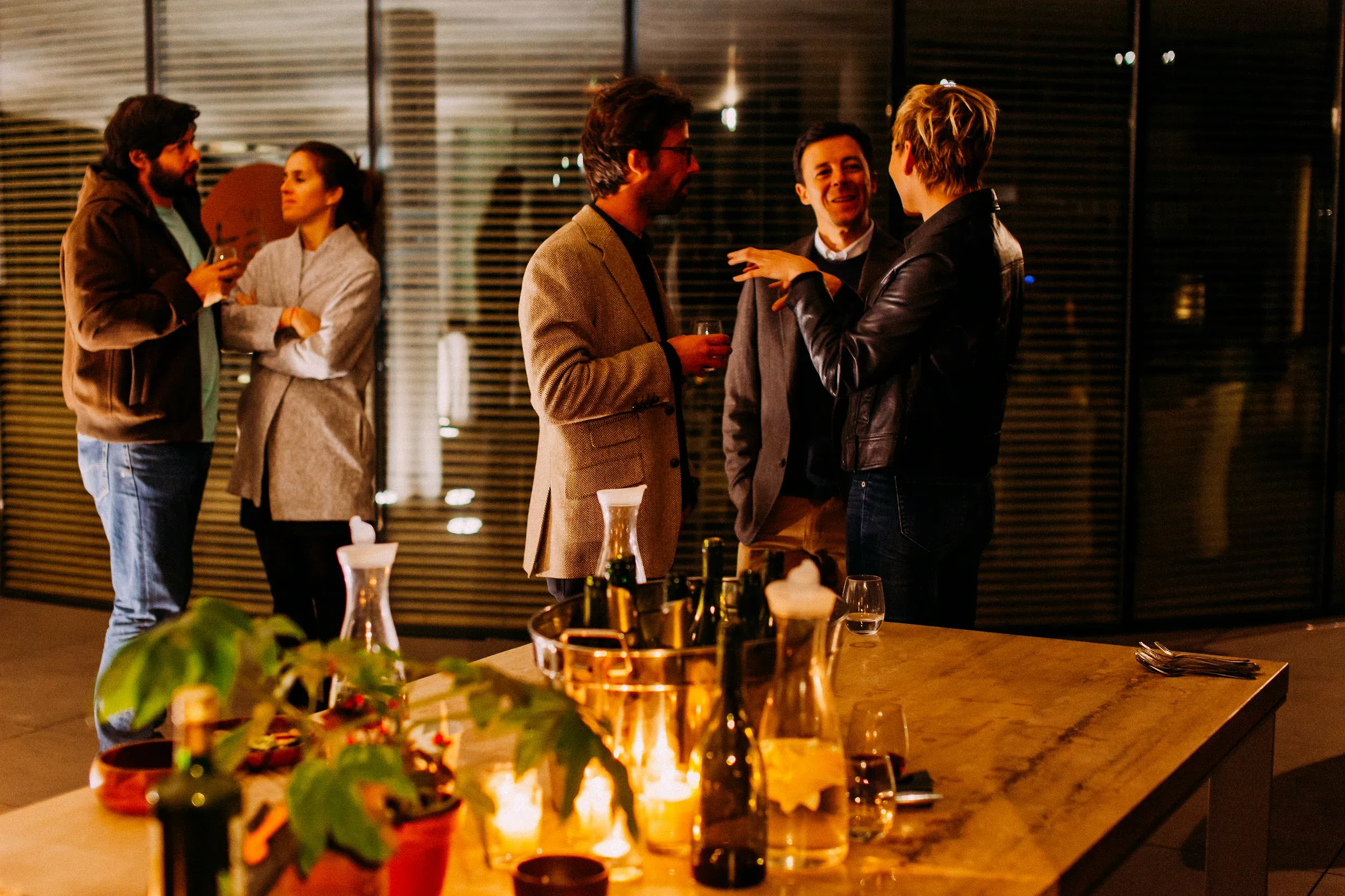 Guests talking in small groups beside a candlelit table of wine at an evening reception
