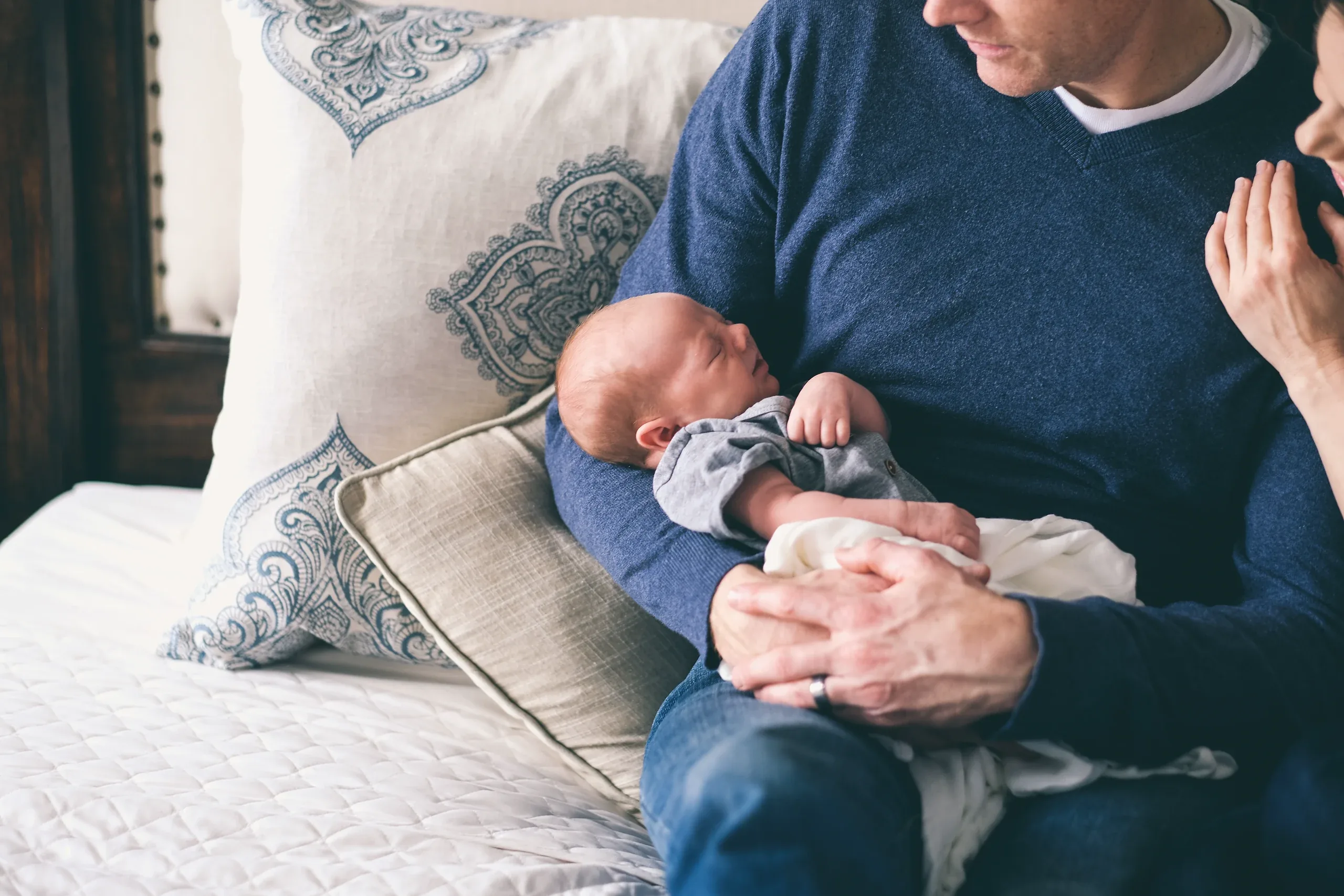 Father on the bed cradling his sleeping newborn against a blue jumper