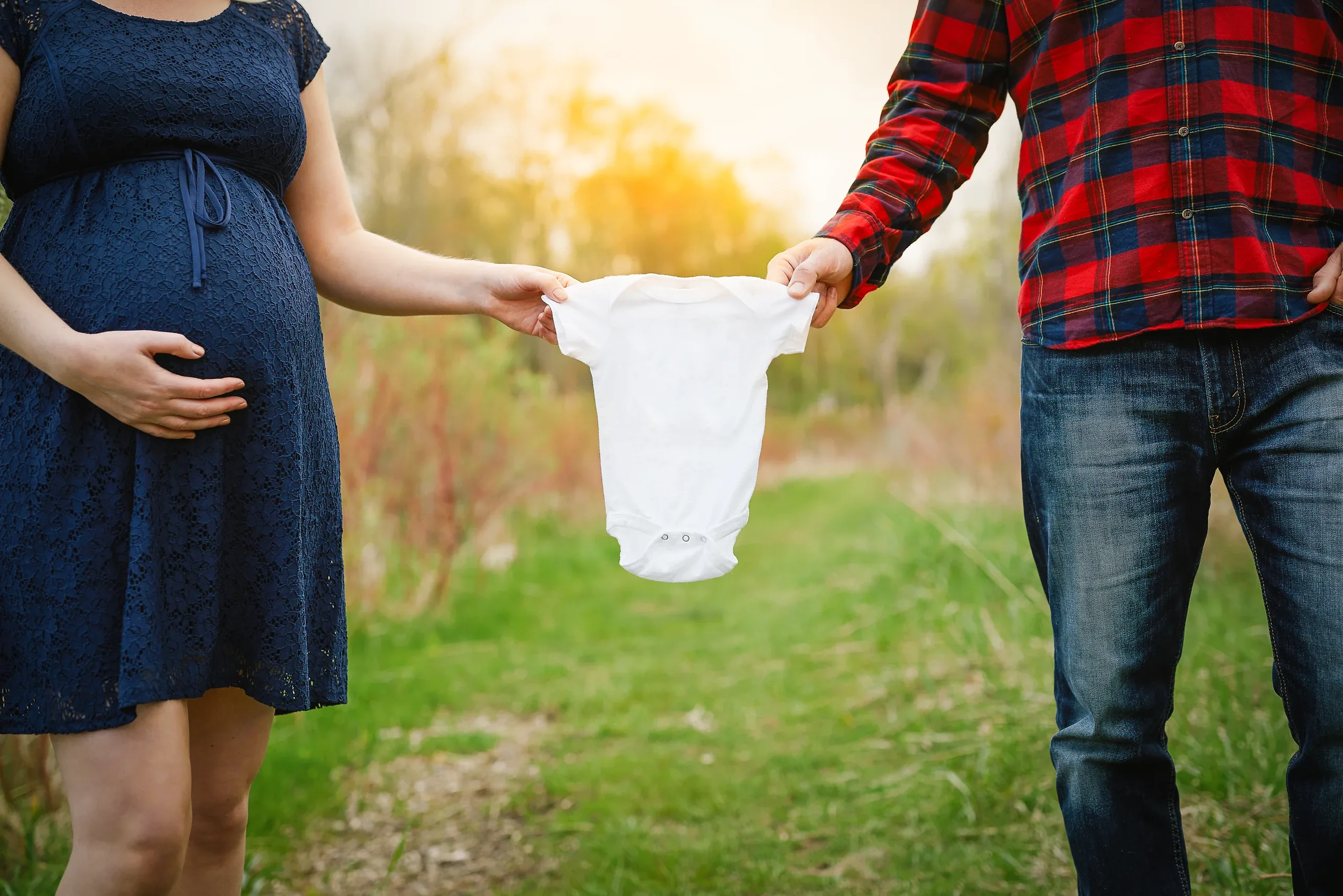 Expecting couple holding a white baby vest between them on a sunlit path