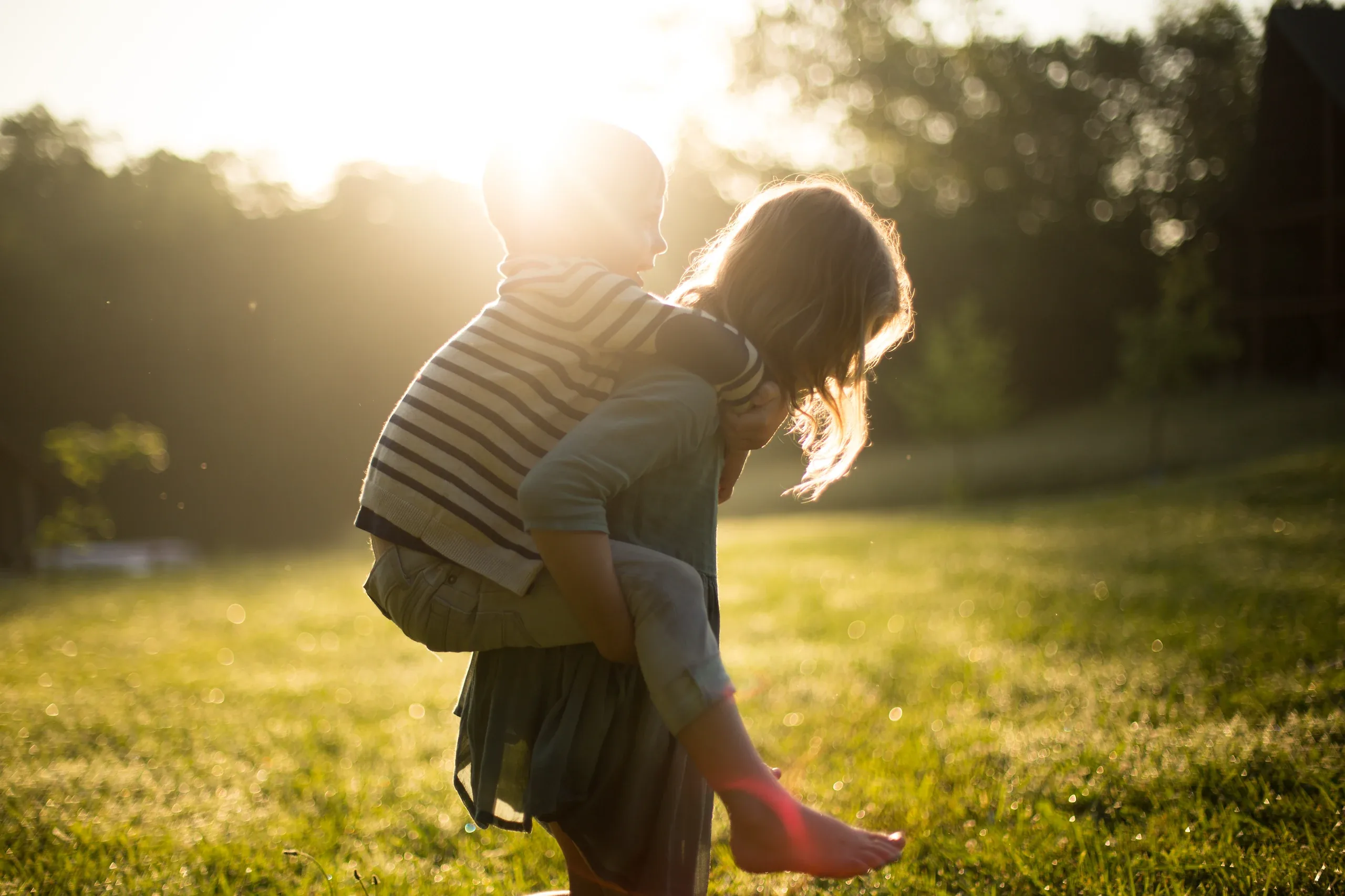 Girl giving her little brother a piggyback across a meadow at sunset