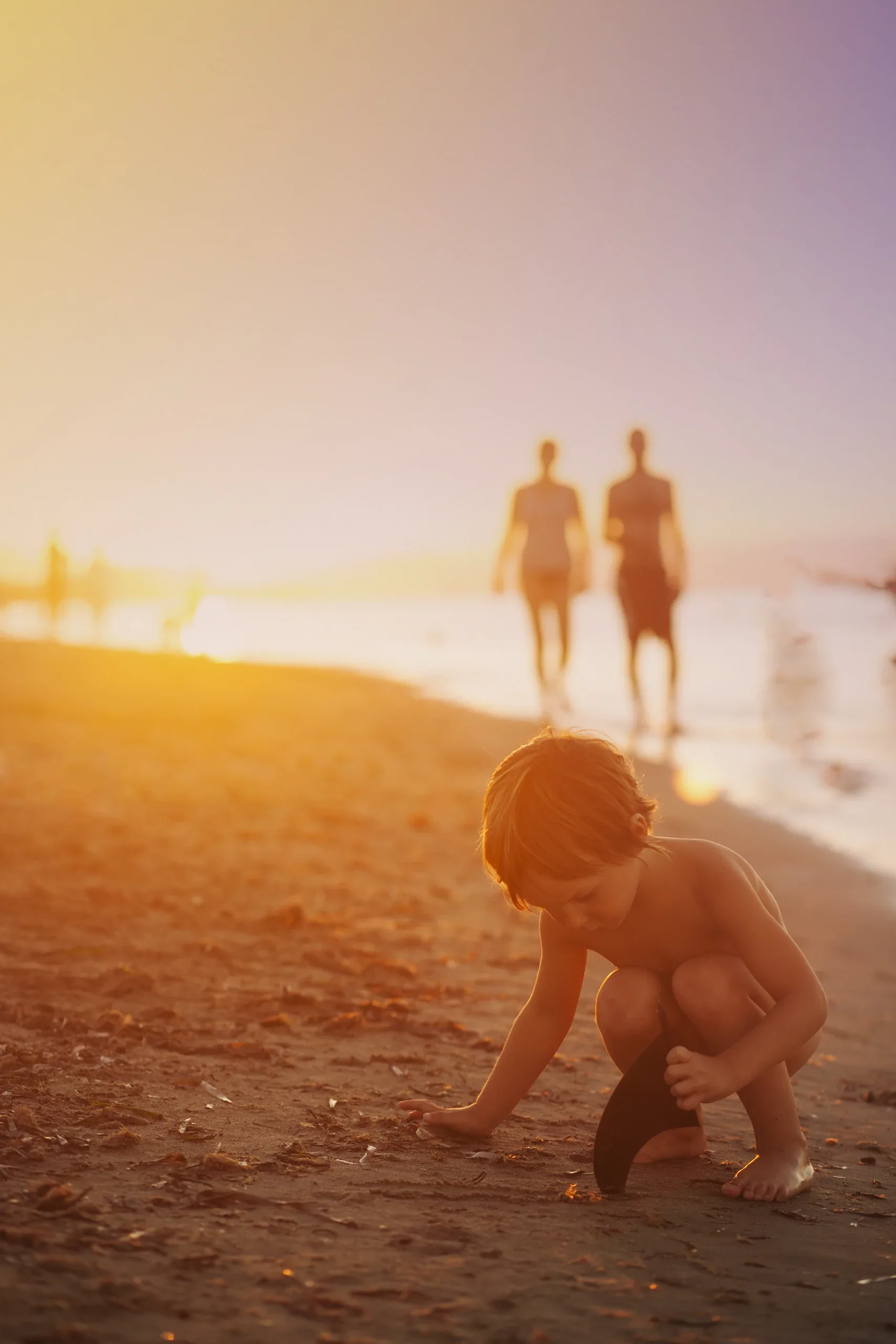 Boy playing in the sand while his parents walk the shoreline at golden hour