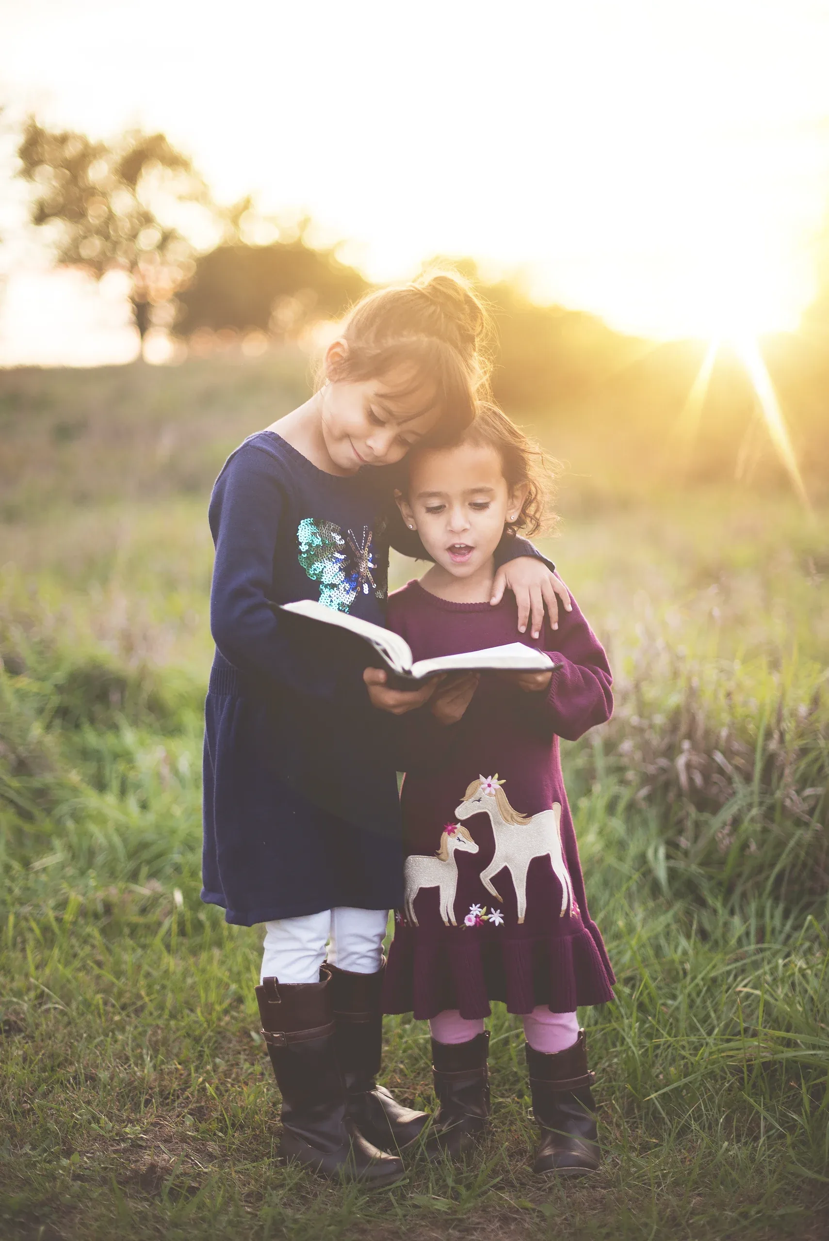 Two sisters reading a book together in a meadow at sunset