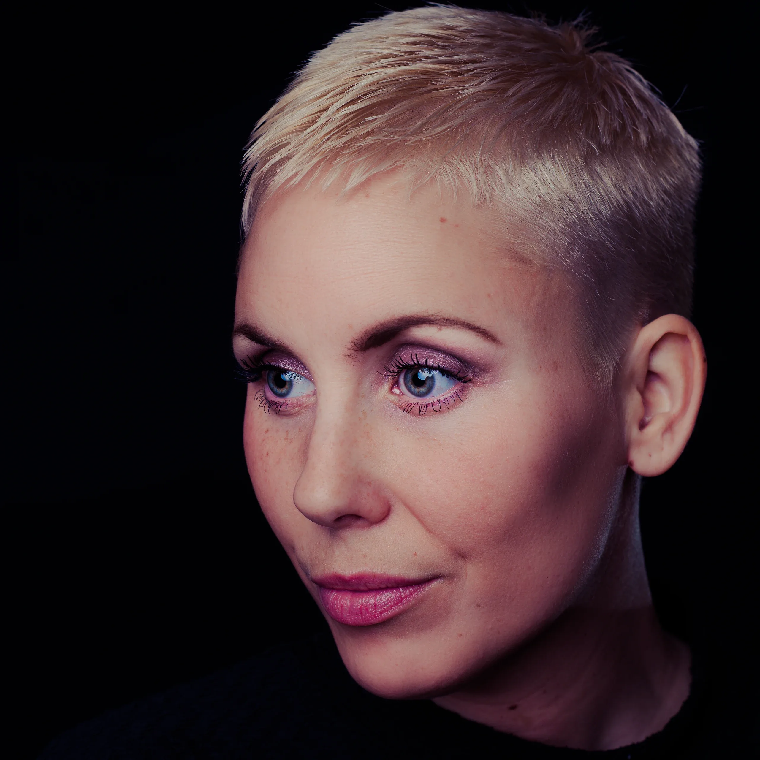 Close studio headshot of a woman with a short platinum crop, lit softly against black