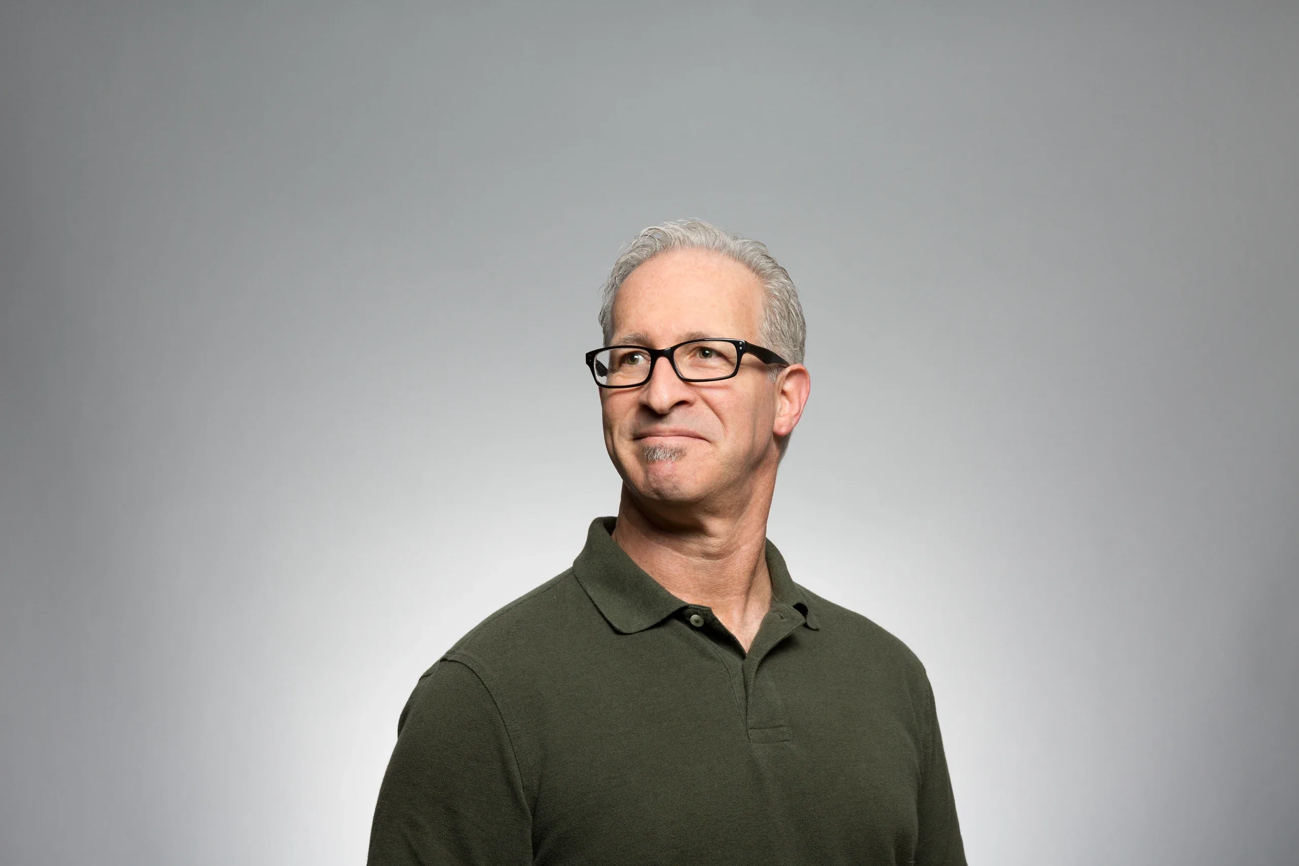 Grey-haired man in glasses and an olive polo shirt against a plain grey studio backdrop