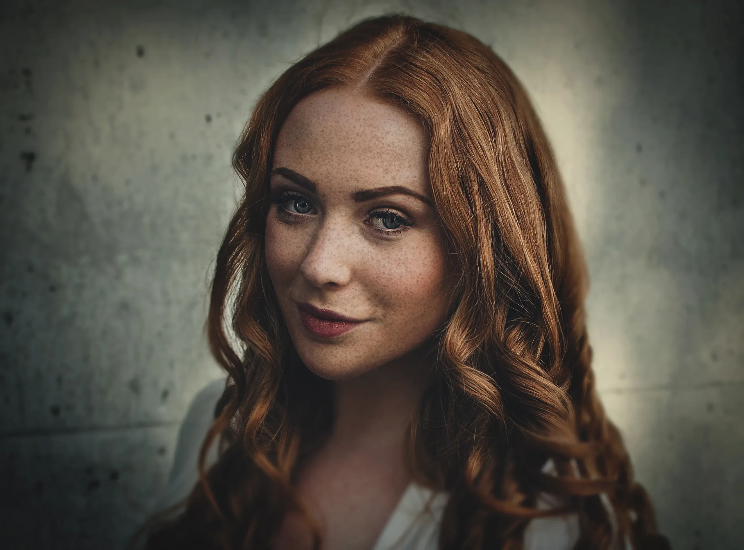 Red-haired woman with long curls and freckles smiling slightly in front of a concrete wall