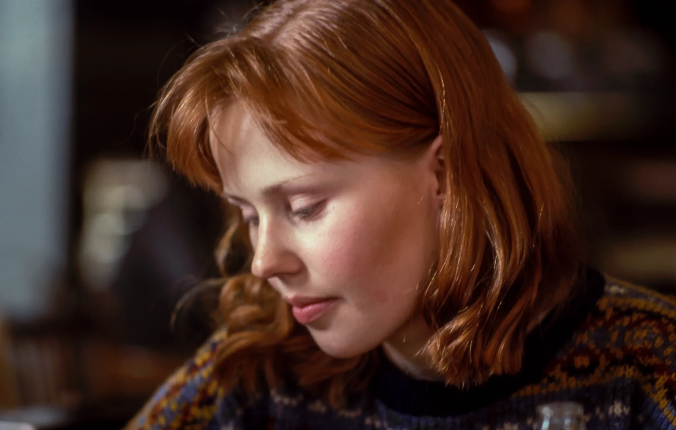 Red-haired woman with a fringe looking down, in warm low light inside a café