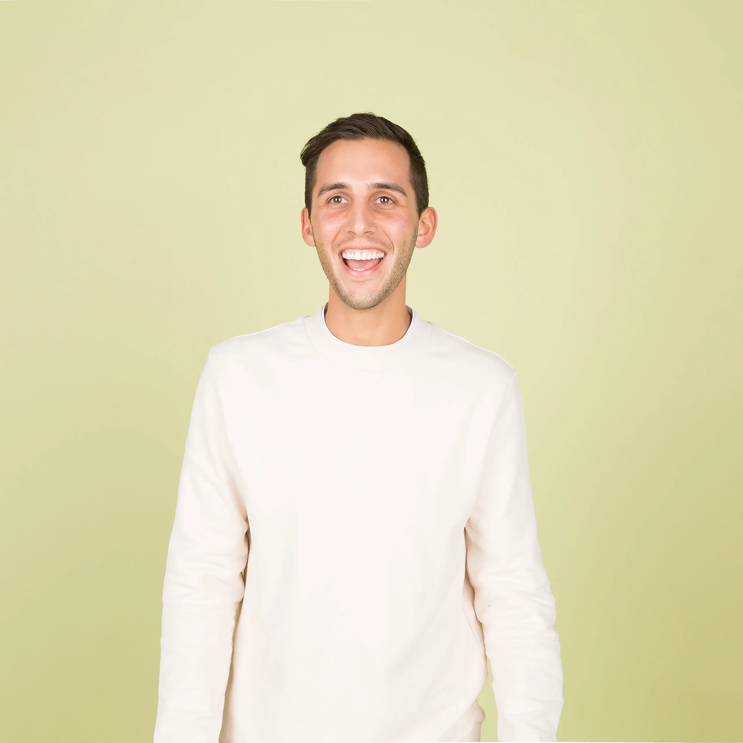 Young man in a cream sweater laughing against a pale green studio backdrop