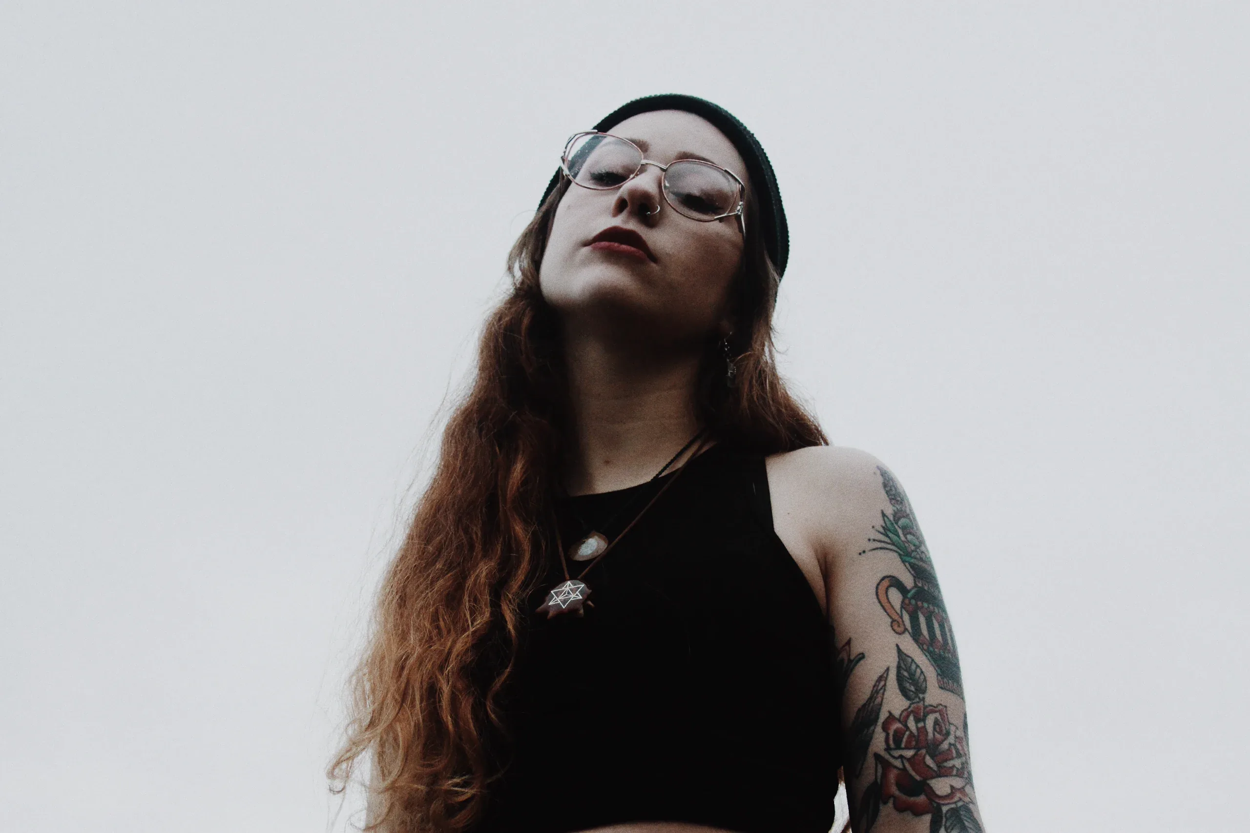 Woman in glasses and a black beanie, tattooed arm, shot from below against a pale sky