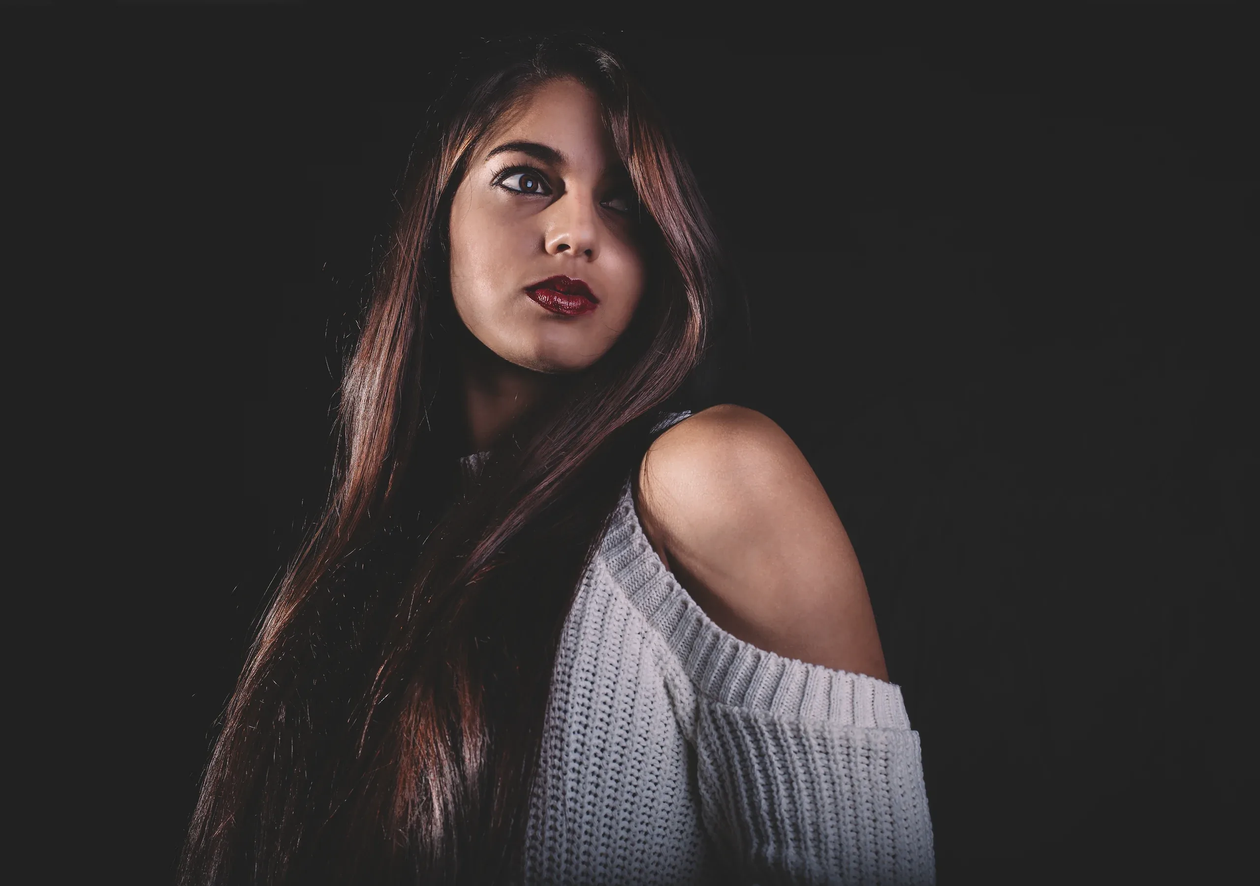 Woman with long dark hair and an off-shoulder sweater lit from the side on black