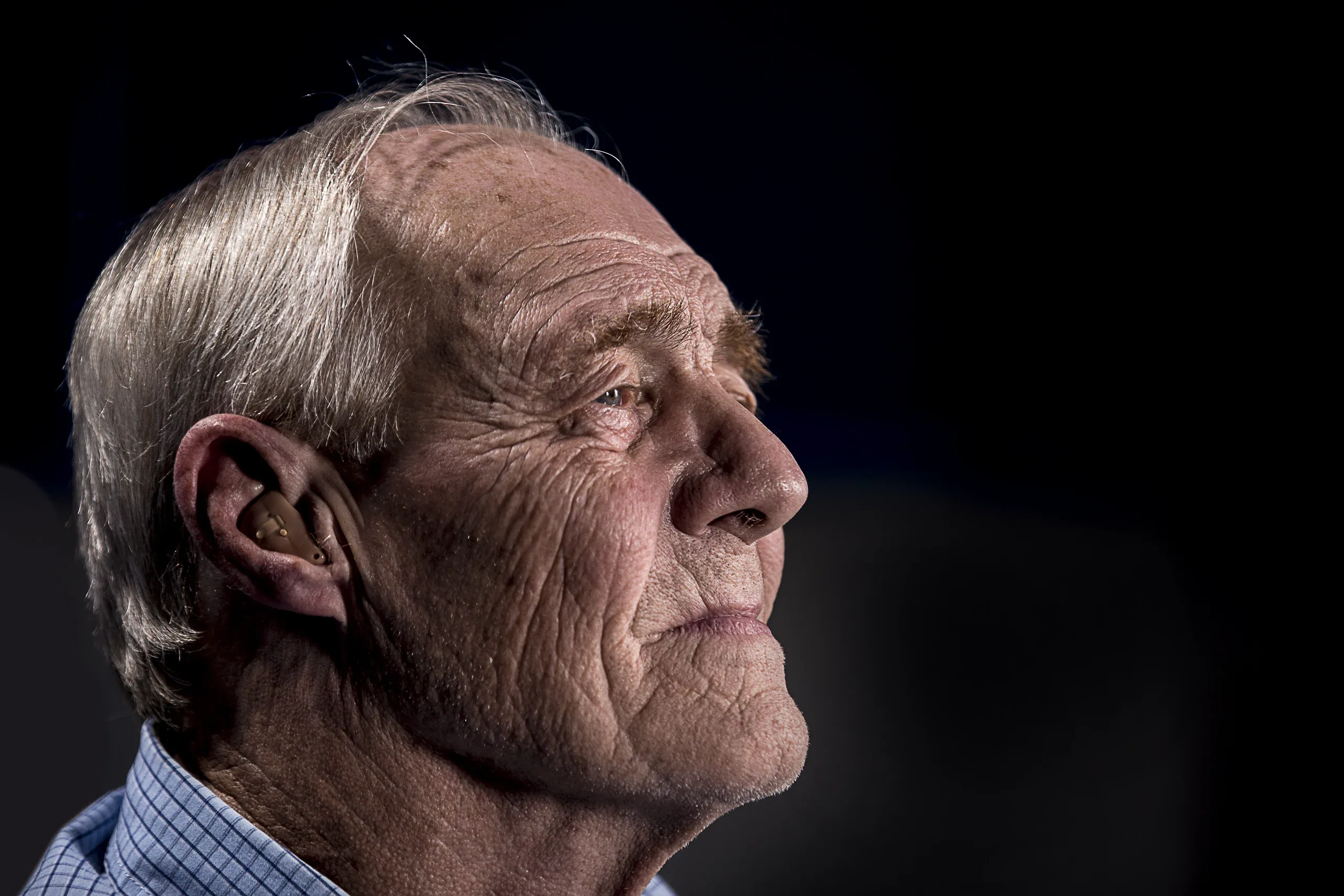 Older man in a checked shirt in profile, his lined face lit from the front against black