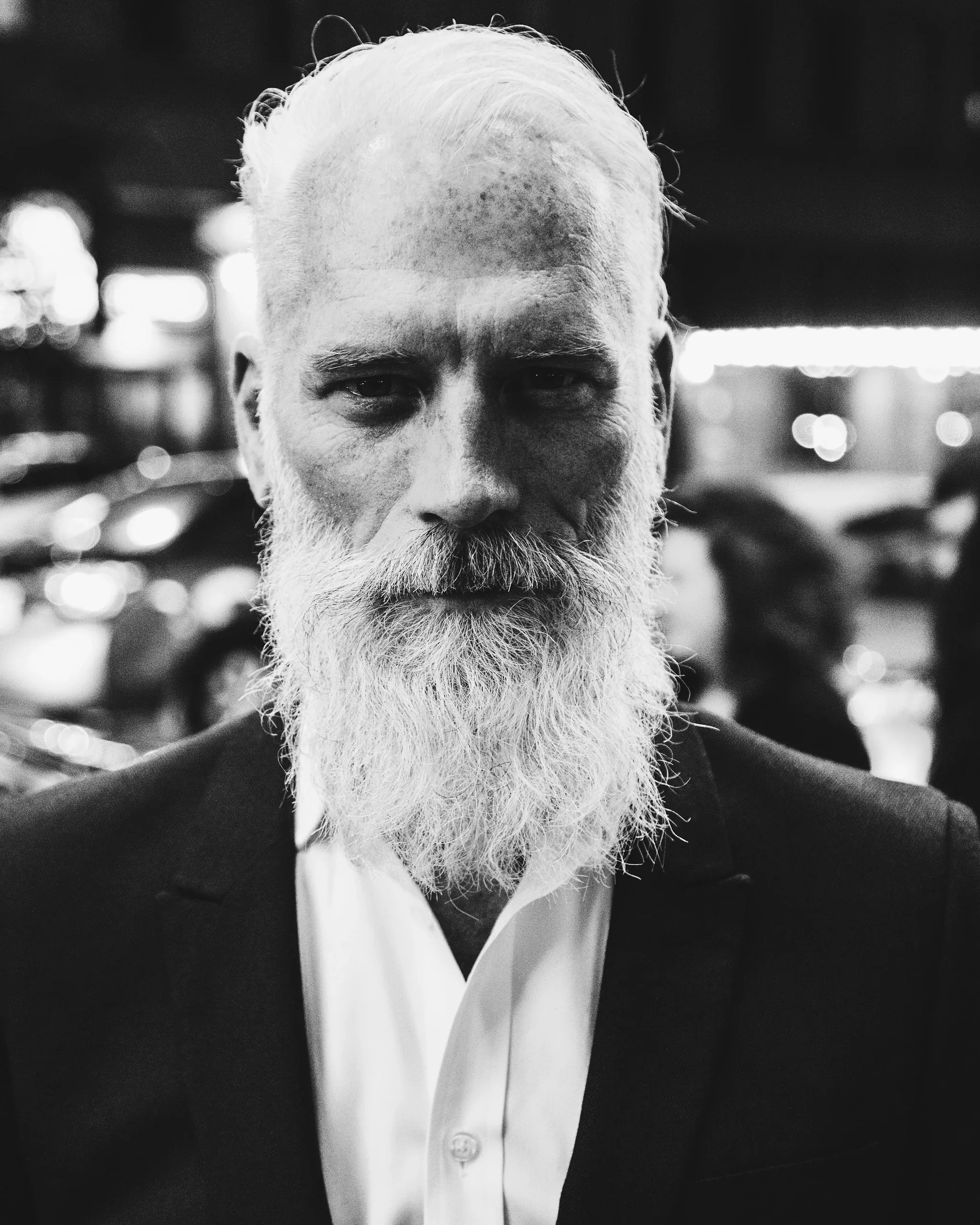 Black and white portrait of a man with a long white beard in a dark jacket on a city street