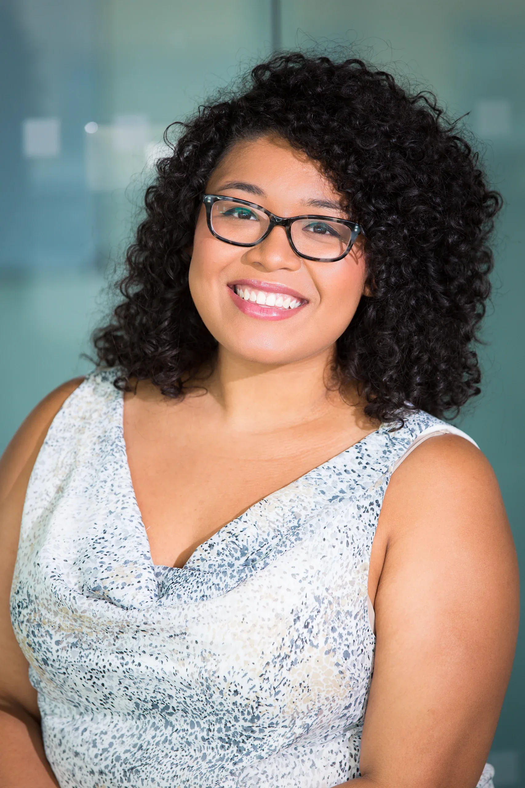 Smiling woman with curly hair and glasses in a patterned top against a teal office wall