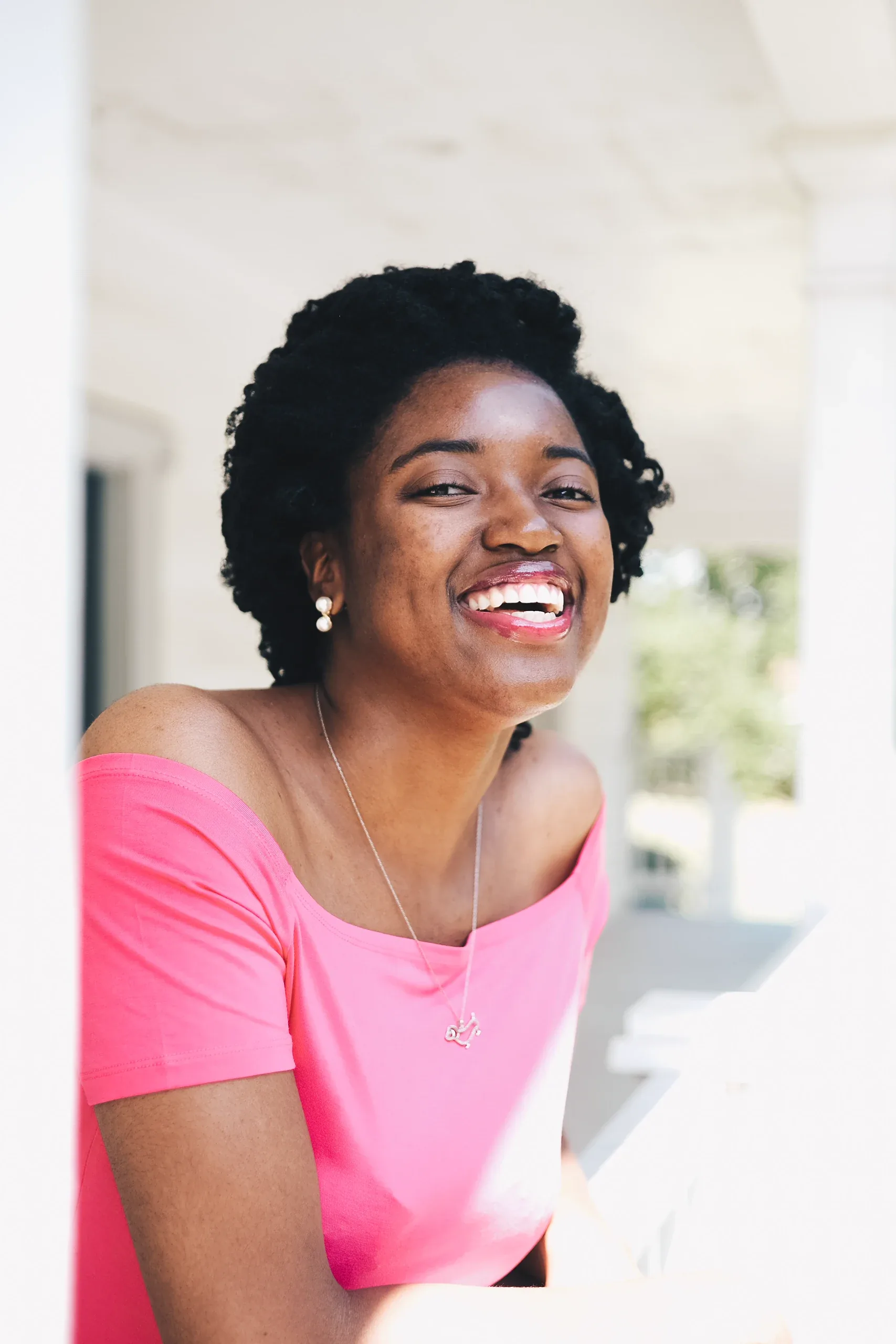 Laughing woman in a pink off-shoulder top leaning on a white porch rail
