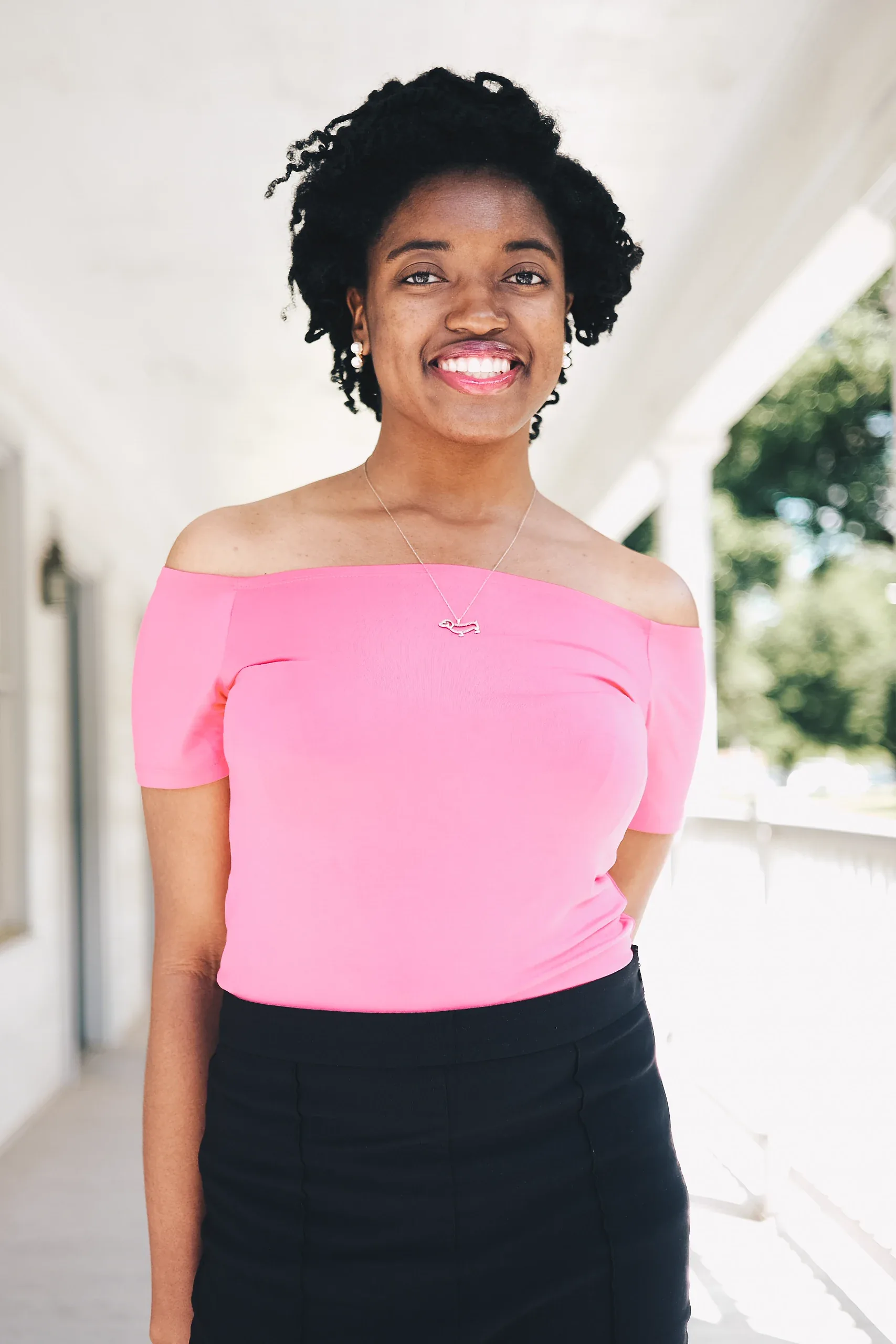 The same woman in the pink top standing full length on the porch, smiling at the camera