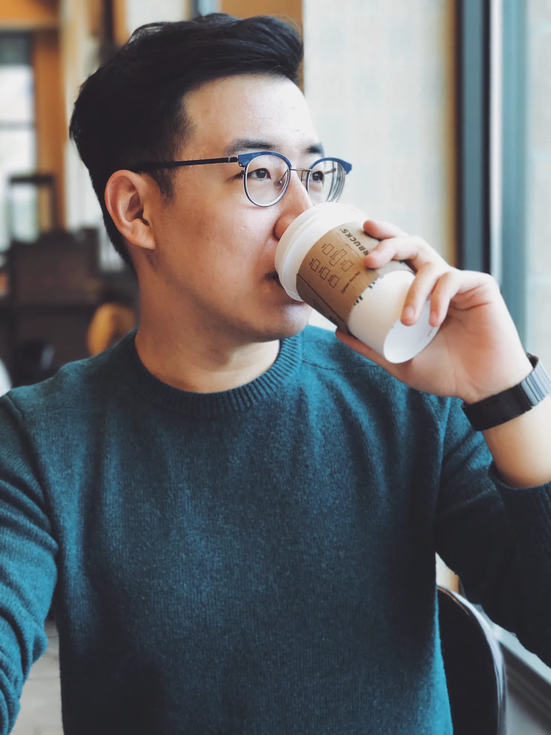Young man in glasses and a teal sweater sipping coffee by a window